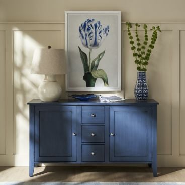 A solid wood blue buffet cabinet with decorative knobs, topped with a white ceramic lamp and a blue vase holding green stems, styled in a classic dining room setting.