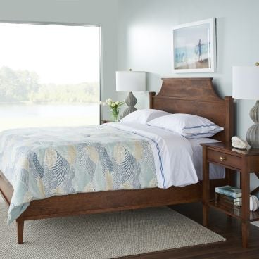 A handcrafted solid wood bed frame with a gracefully curved headboard and matching wooden nightstand in a bright, serene bedroom overlooking a scenic lake.