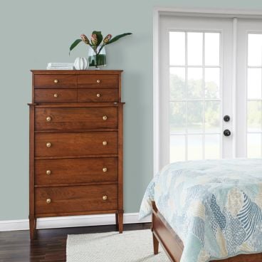 Solid wood highboy dresser with six drawers and brass hardware, positioned beside a bed in a serene bedroom with natural light.