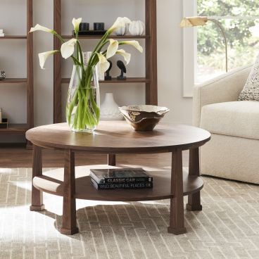 Round wooden coffee table crafted in a classic style, featuring a lower shelf for storage, displayed with a vase of white flowers in a modern living room setting
