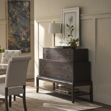A stylish dark wooden chest of drawers with brass handles, elegantly displayed in a sunlit room, accompanied by artwork and decorative items on top.