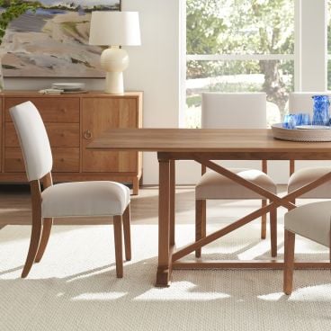 Elegant dining area showcasing a wooden table with crossbeam support and matching chairs, set against a backdrop of a wooden sideboard and large windows.