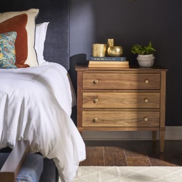 Solid wood three drawer nightstand with brass knobs beside a bed, styled with books, gold vases, and a plant against a dark bedroom wall.