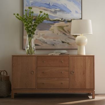 Contemporary solid wood storage consule with brass hardware, topped with a stylish lamp and a vase of fresh greenery, framed by a large abstract artwork