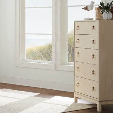 Solid wood five-drawer dresser with gold ring pulls, set against a bright coastal backdrop.