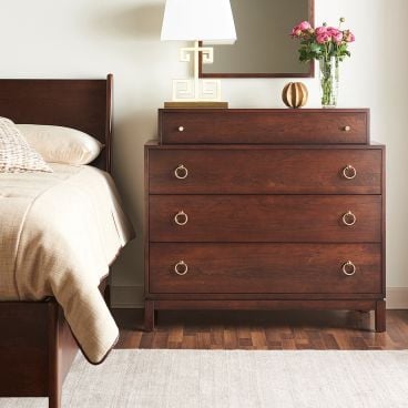 Solid wood four-drawer dresser with gold ring pulls, styled with a lamp, mirror, and vase of flowers beside a matching bed.