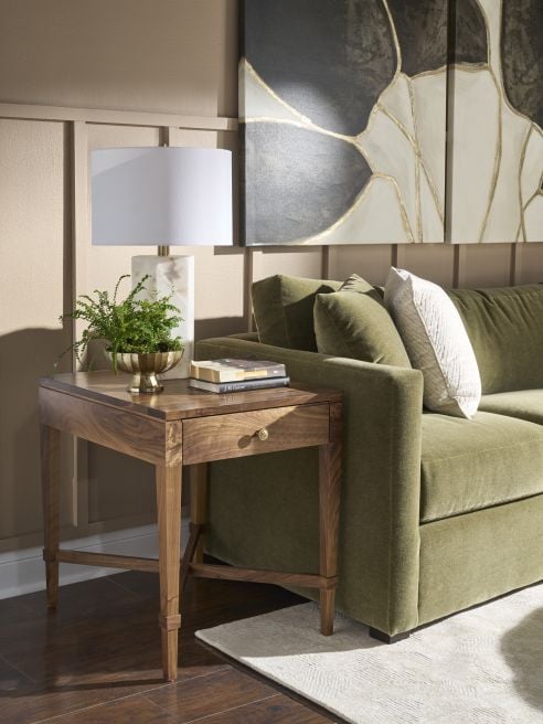 Solid wood end table with drawer beside a green sofa, styled with a white marble lamp, books, and a potted fern on warm neutral walls.