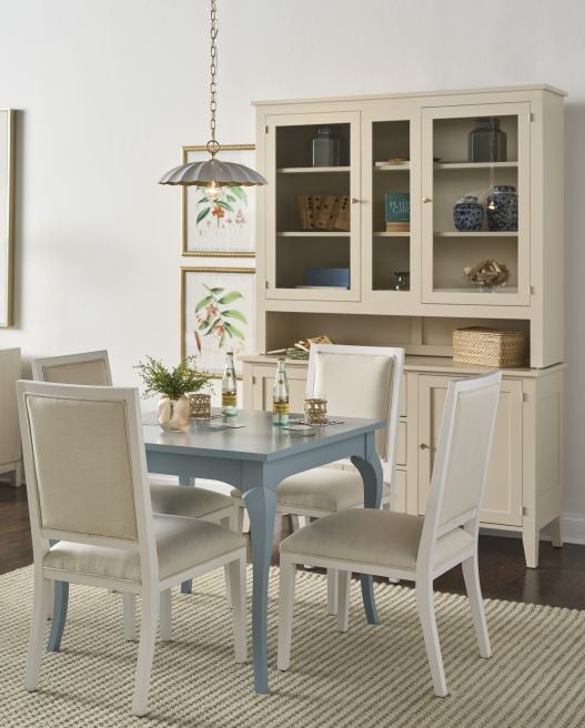 Blue painted solid wood game table with four upholstered chairs, paired with a cream glass door hutch in a bright dining room.