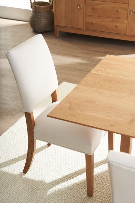 A close-up of a dining chair with a simple, modern design featuring a light wood frame and a padded, upholstered seat in a soft cream color