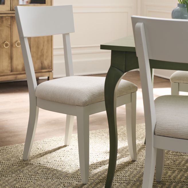 Light upholstered dining chairs at a table on a woven rug with a wood cabinet and vase of tulips in the background.