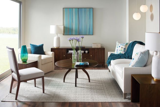 Bright and modern living room featuring a round cocktail table with sculptural legs, sleek sideboard with storage compartments, tailored white seating, minimalist side tables, and layered accent pillows and throws for a cozy yet sophisticated aesthetic.