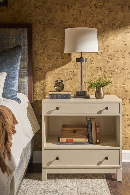 Modern painted wood nightstand next to a bed, featuring stacked books, a vintage clock, metal lamp, and small plant on top.