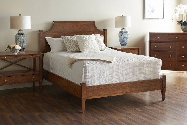 Cherry wood bed with a curved headboard, complemented by matching nightstands and a dresser, in a serene bedroom with a white quilt and blue ceramic lamps.