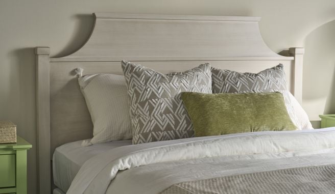 Close up of a shaped hardwood bed headboard framed by patterned euro shams and a green lumbar pillow.