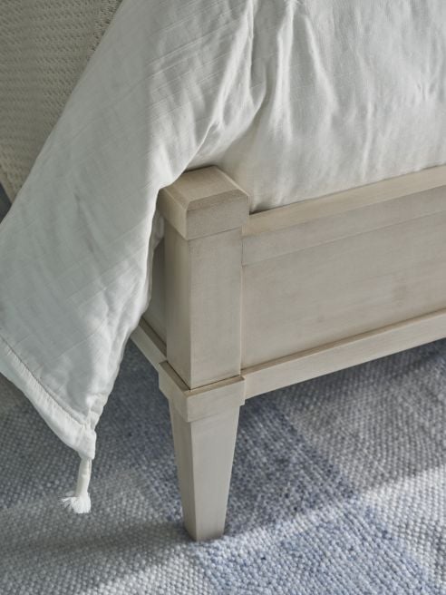 Corner detail of a painted solid wood bed footboard with tapered leg, shown with white bedding on a blue woven rug.