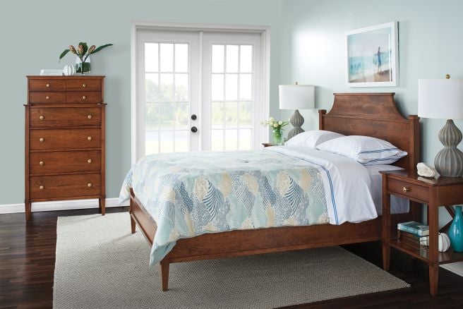 Cherry wood bed with a curved headboard, matching nightstands, and a tall chest of drawers, styled in a bedroom with French doors and serene lake views.