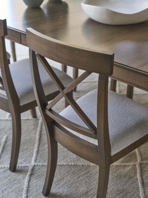 Detail view of a hardwood chair showing curved legs, cross back design, and textured neutral seat cushion at a dining table.