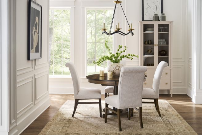 Appalachian hardwood pedestal table and fabric covered dining chairs on a patterned rug, with large windows and storage cabinet in the background.