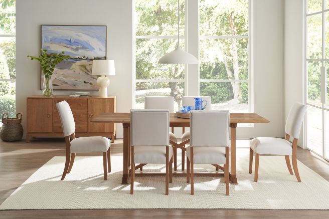 A bright dining room featuring a wooden dining table and chairs set against large windows, complemented by a wooden sideboard and neutral decor