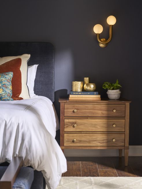 Hardwood bedside chest with three drawers on warm wood floors, shown next to an upholstered bed and modern brass wall sconce.