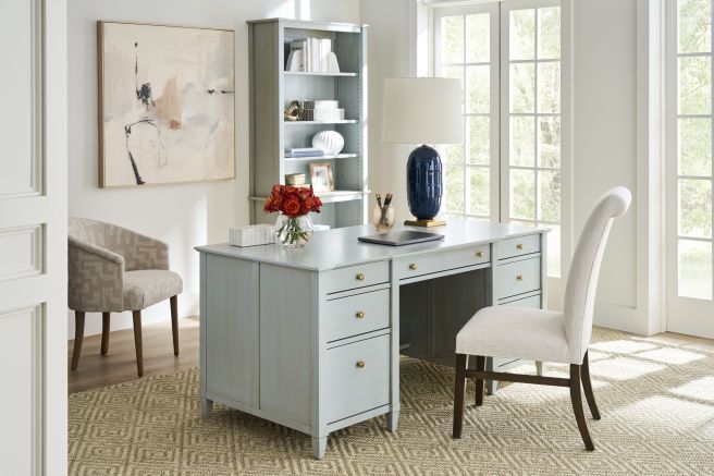Solid wood writing desk with drawers and laptop, paired with a high back upholstered chair and shelving unit in a light filled office.