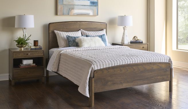 Solid wood bed with rounded headboard and matching nightstands, styled with neutral quilted bedding and blue accent pillows in a bright bedroom.