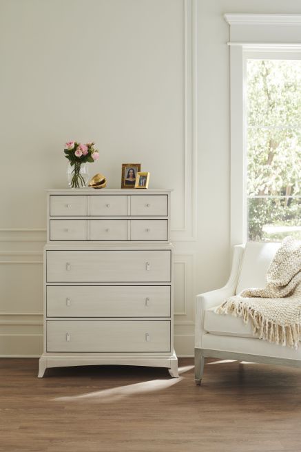 A tall, handcrafted solid wood highboy dresser painted white featuring a combination of small and large drawers, styled in a bright bedroom next to a cozy armchair and large window.