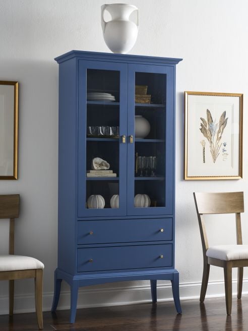 Tall hardwood display cabinet in a deep blue painted finish, featuring glass doors, storage drawers, and brass knobs in a modern dining room.